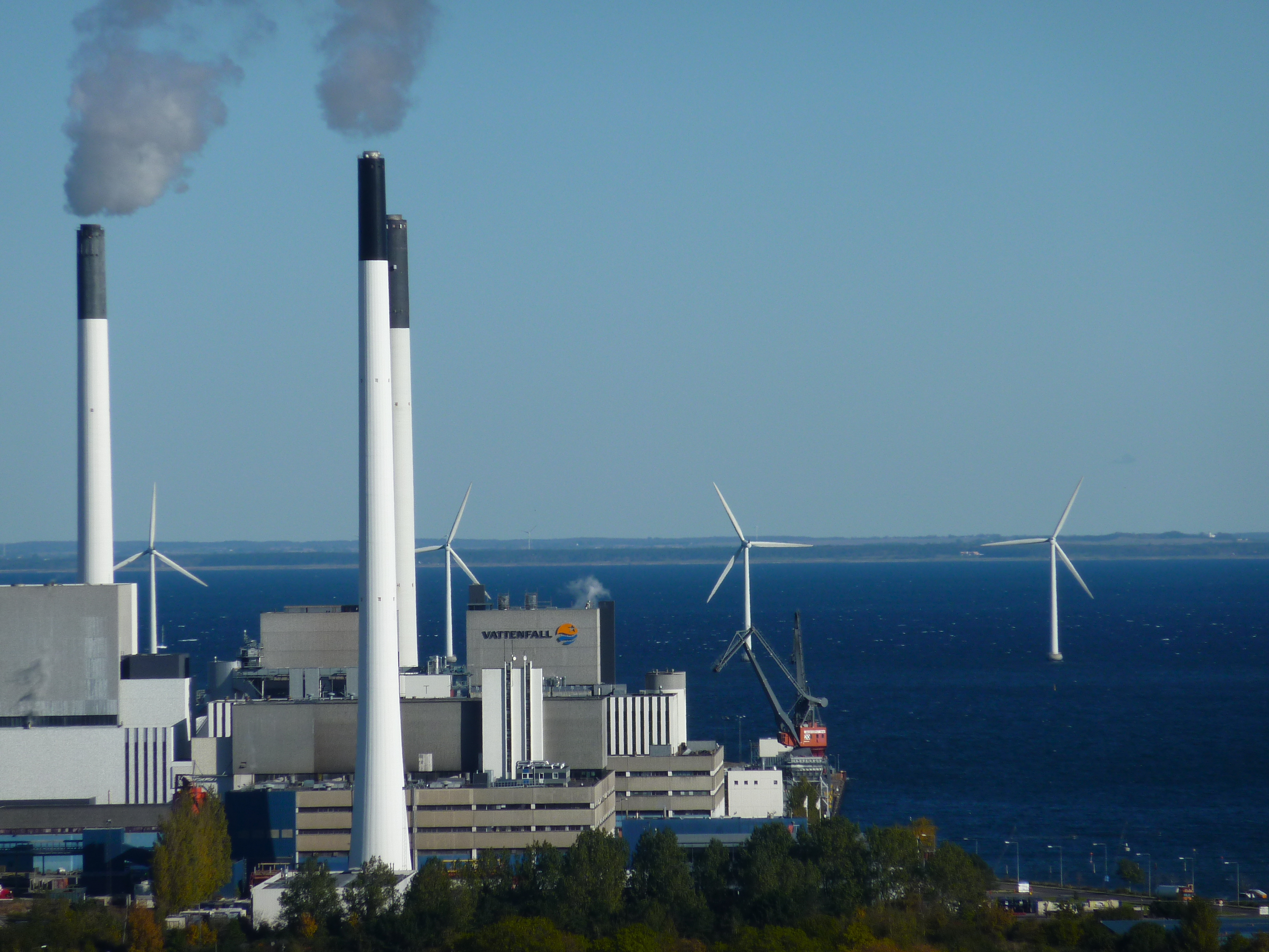 white smoke stacks near a bay, 4 windmills sit in the bay