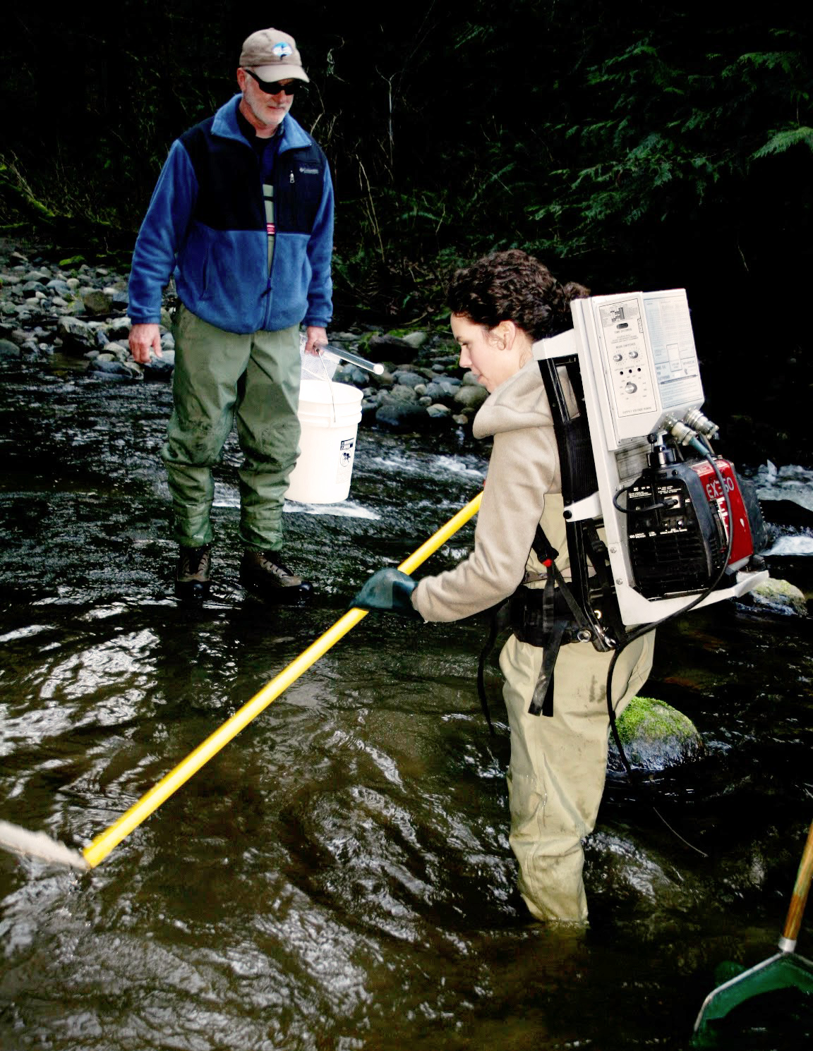 A student wearing a large, rectangular contraption on her back containing pumps holds a yellow pole to a stream while  professor observes.