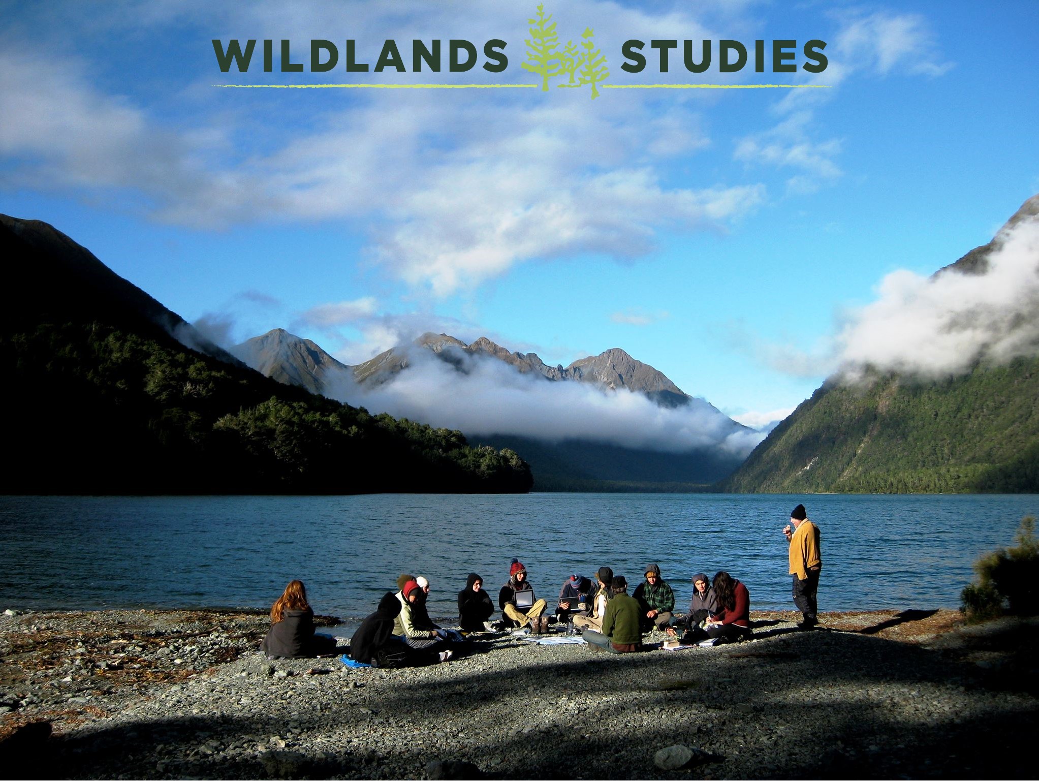 Students sitting in front of a lake with mountains and low hanging clouds in the background. The text &quot;Wildland Studies&quot; appears at the top.