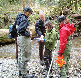 Four students gather around a professor near a stream. The professor is pointing to a book one of the students is holding