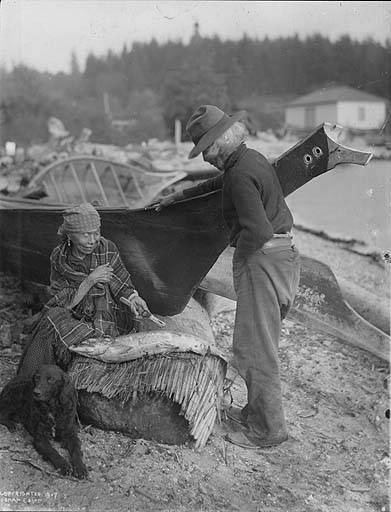 Snohomish man and woman pose with salmon by canoes, Tulalip Indian Reservation, Washington, 1907