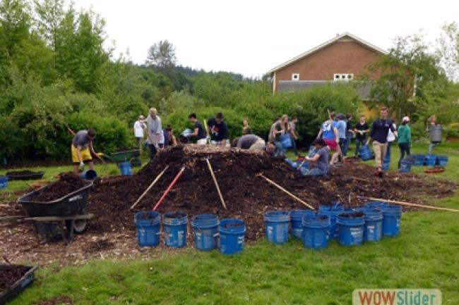 A large group of Western students work near a 6' tall pile of dirt. Surrounding the freshly moved dirt pile are a dozen rakes and 10 blue buckets.