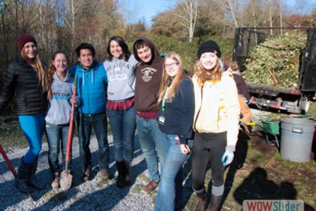 A group of 7 Western students standing near the rear of a truck filled with grass clippings and plant debris. A rake, shovels and hand tools are laying on the ground nearby.
