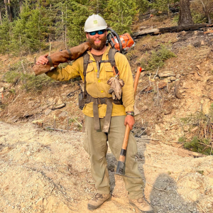 The speaker standing in a forest with wildland firefighting equipment 