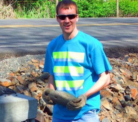 Portrait of Jonnel Deacon, holding a rock and standing in front of a road, wearing sunglasses and a bright blue t-shirt with reflective yellow stripes