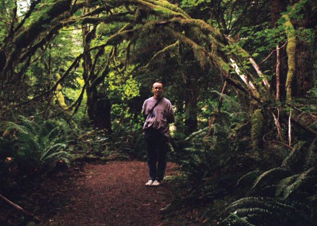 Portrait of Bo Alfus surrounded by trees deep in the woods, wearing an oversized purple t-shirt