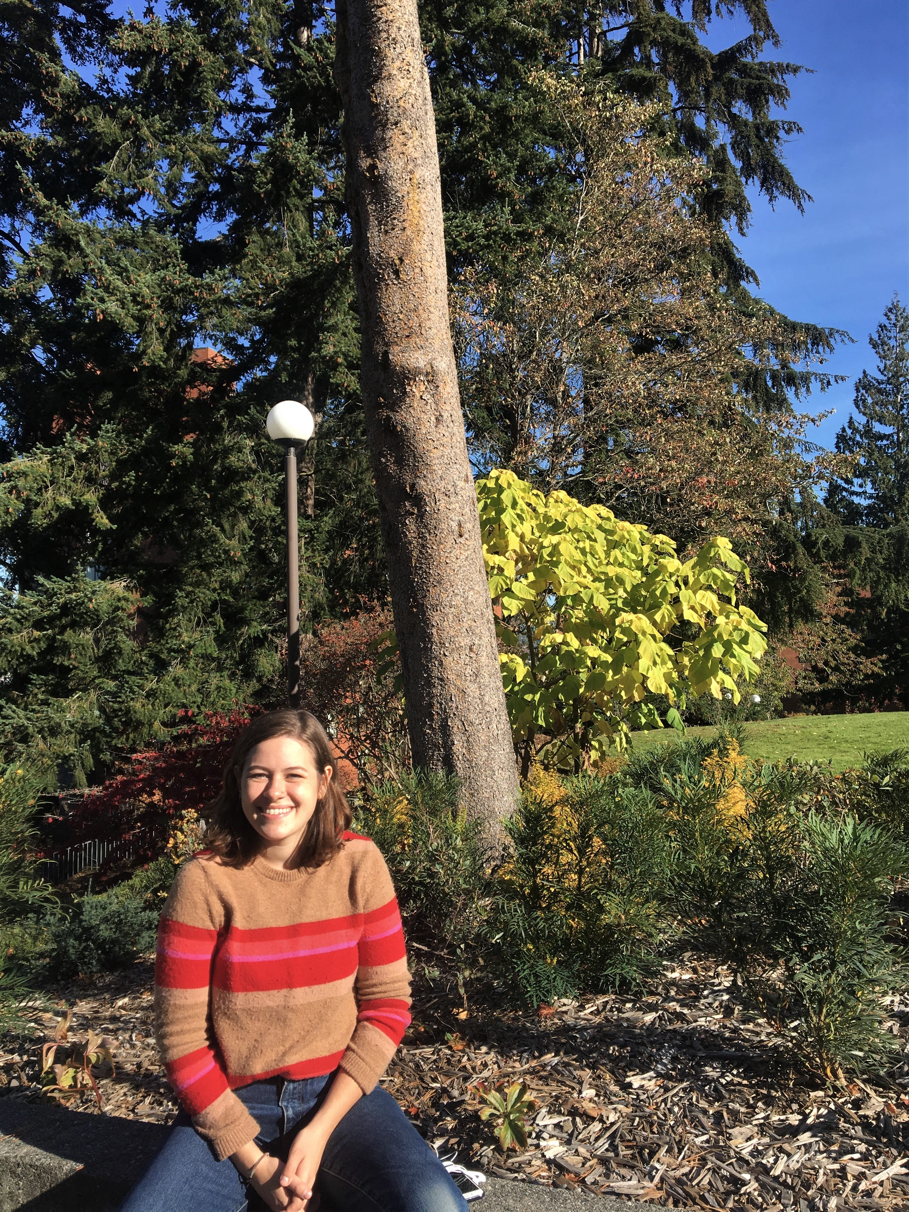 Portrait of Alyssa Webster sitting in front of a tree and lawn, wearing a camel-colored sweater with 2 sets of red, orange, pink, red, and beige stripes (of varying width) and blue jeans