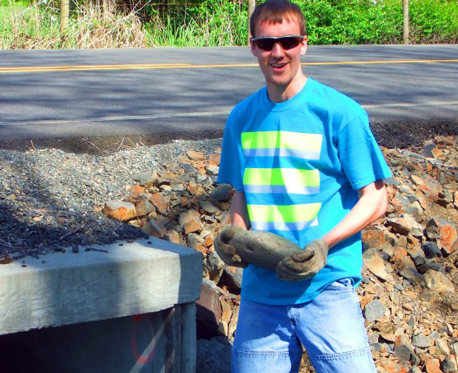 Portrait of Jonnel Deacon, holding a rock and standing in front of a road, wearing sunglasses and a bright blue t-shirt with reflective yellow stripes
