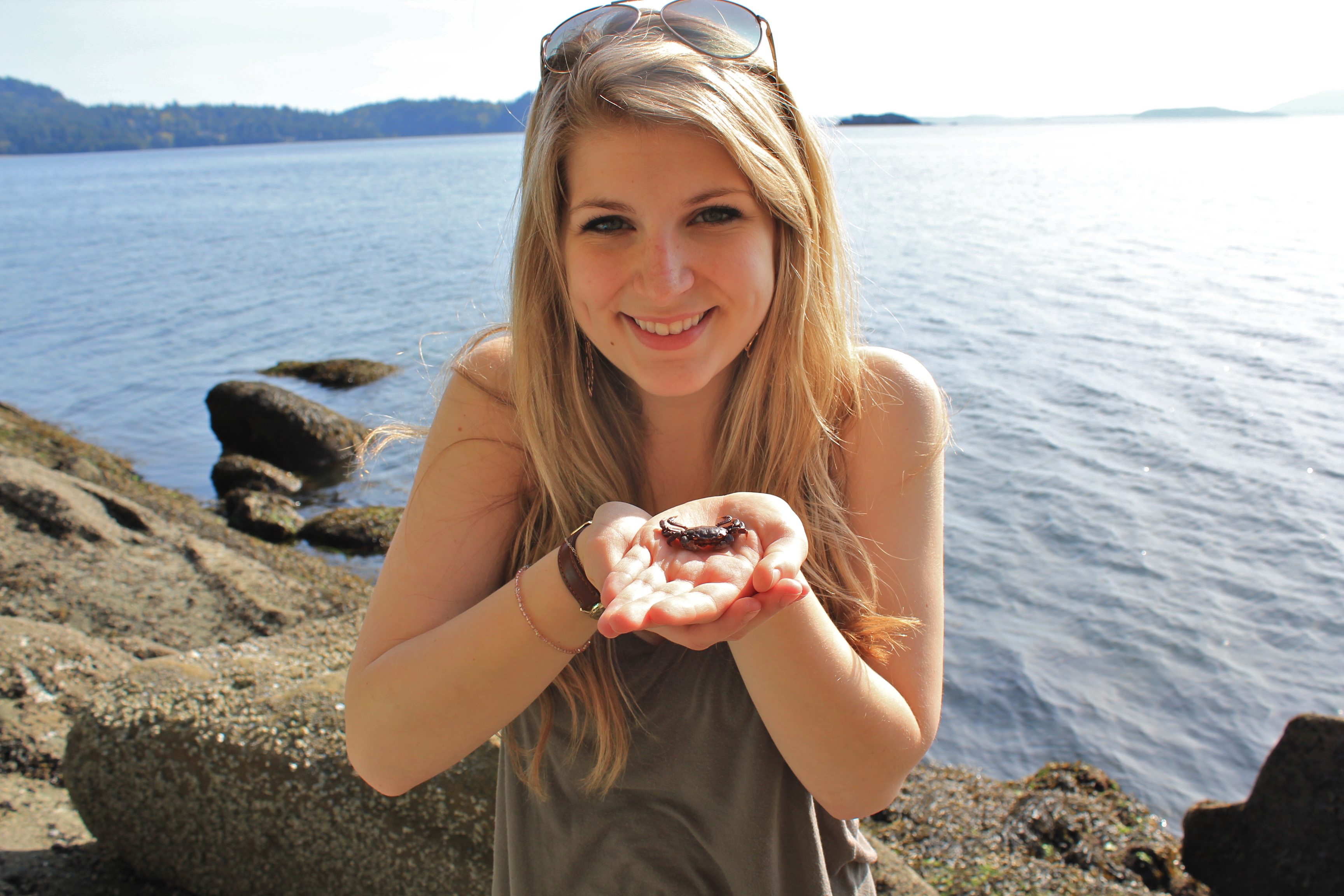 Portraint of Christine Hanley standing on a sunny beach and smiling, with hands outstretched towards the camera presenting a small crab