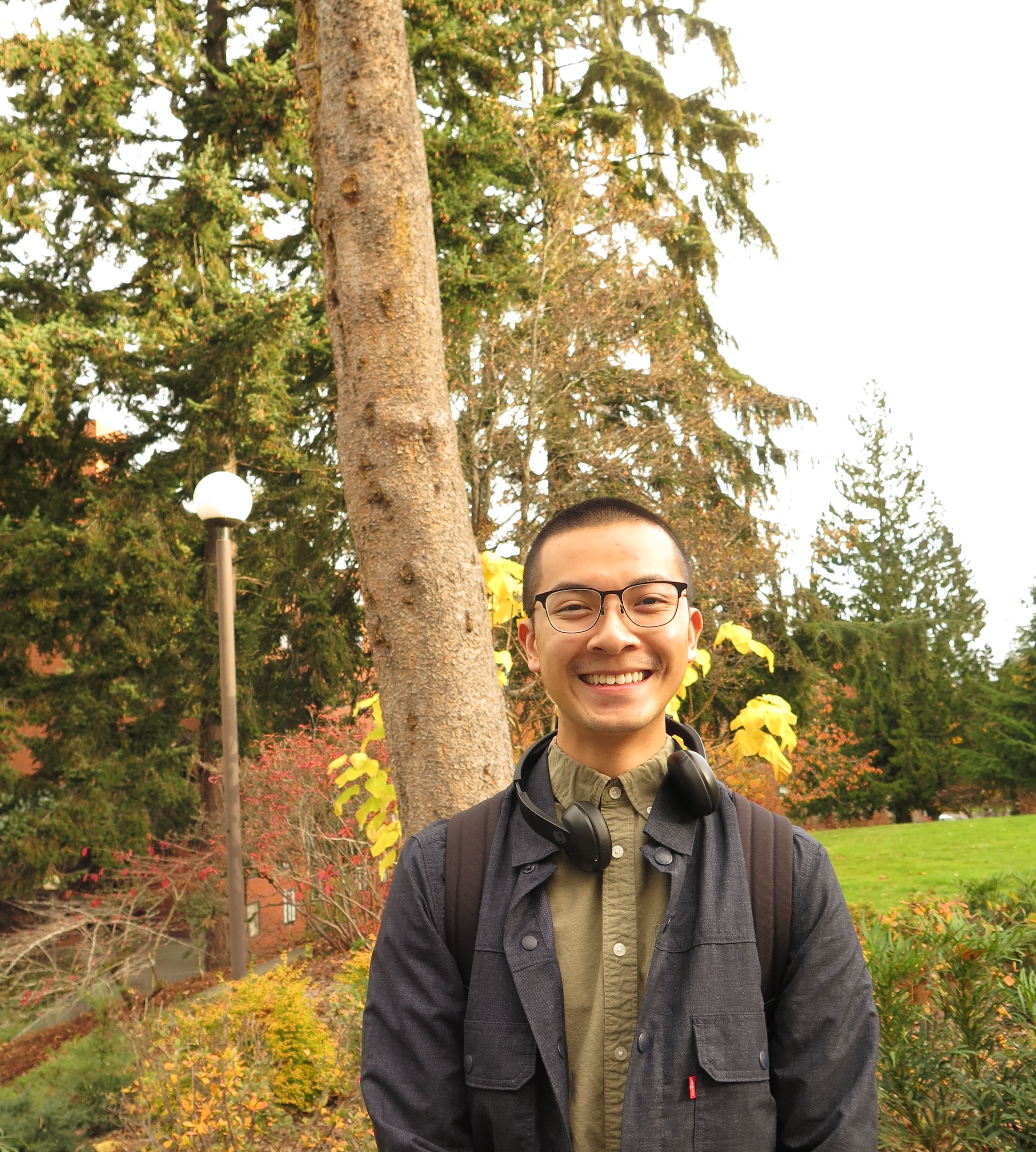 Portrait of Peter Cung standing in front of a tree and lawn, wearing a green button-down shirt, Levi&#039;s trucker jacket, black bluetooth headphones, black metal glasses, and black backpack