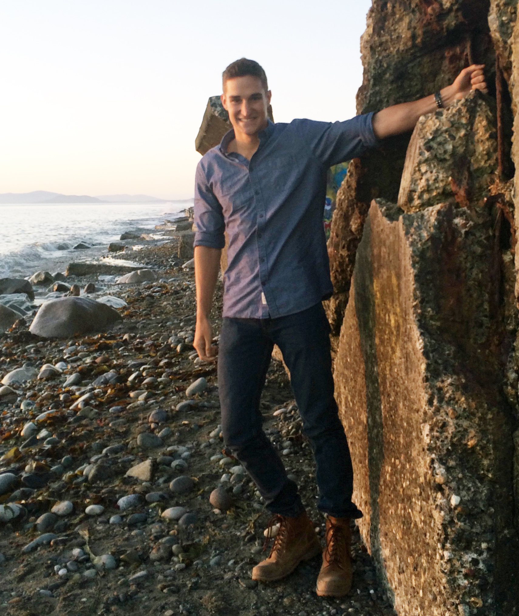 Portrait of Sam Schultz on a rocky beach close to sunset, hanging off of a boulder wearing a blue button up shirt