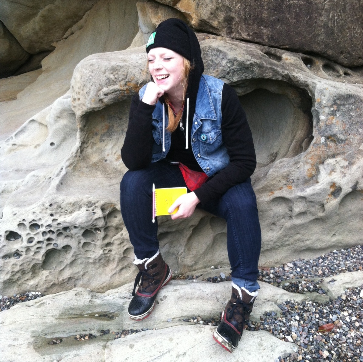 Portrait of Deven Vilar sitting on a concavity in sandstone, wearing a jean vest over a black hoodie, a black beanie, and black converse