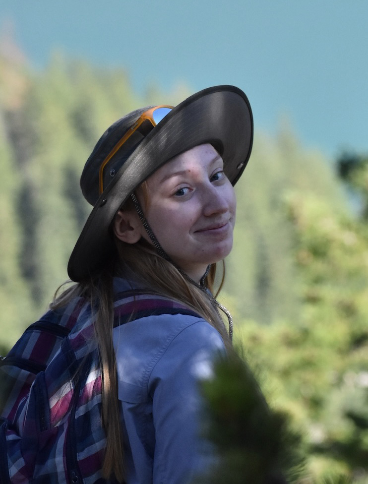 Brianna Benner is outside wearing a backpack and hat looking back over her shoulder.