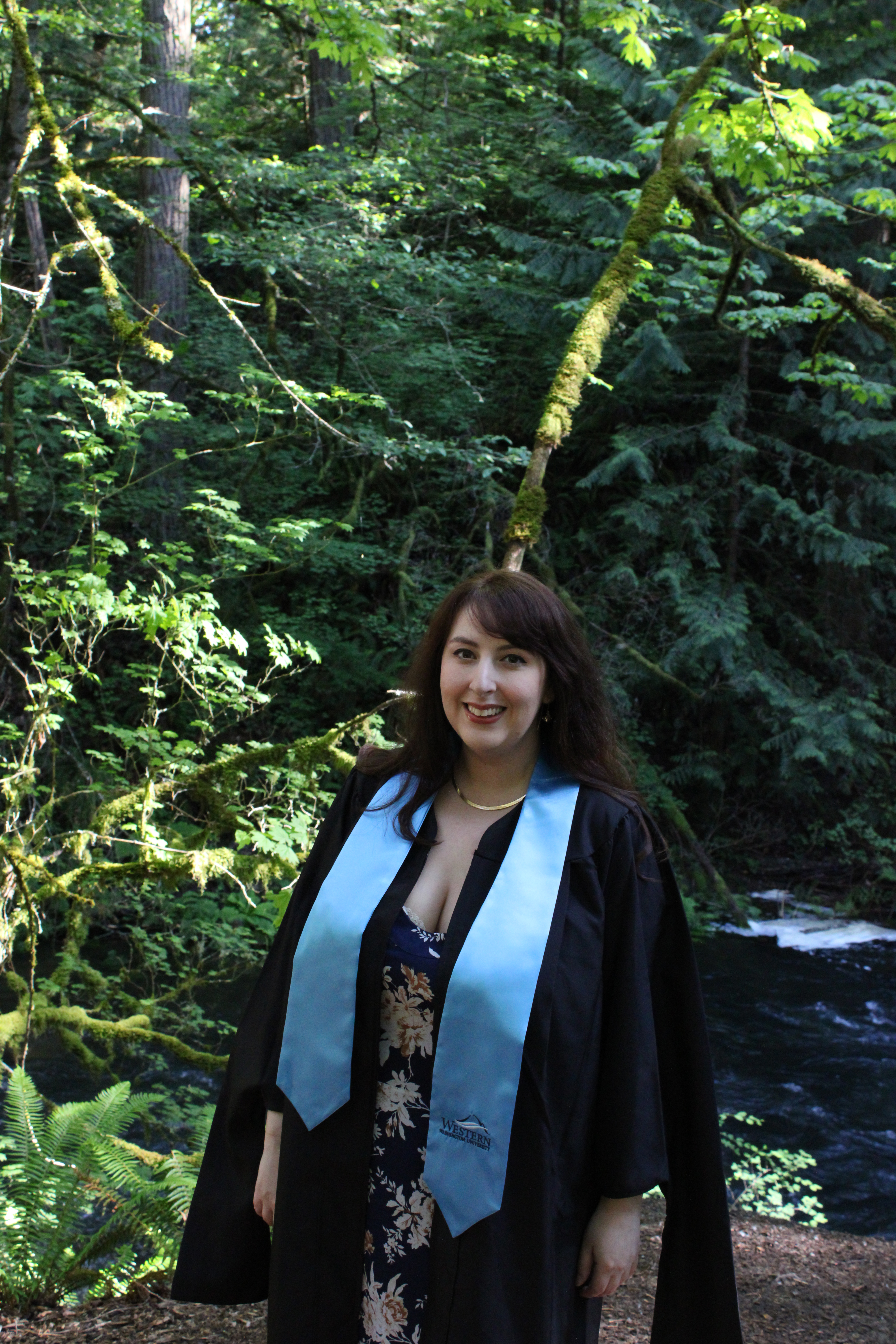 Holly Suther poses in a shady forest wearing graduation regalia