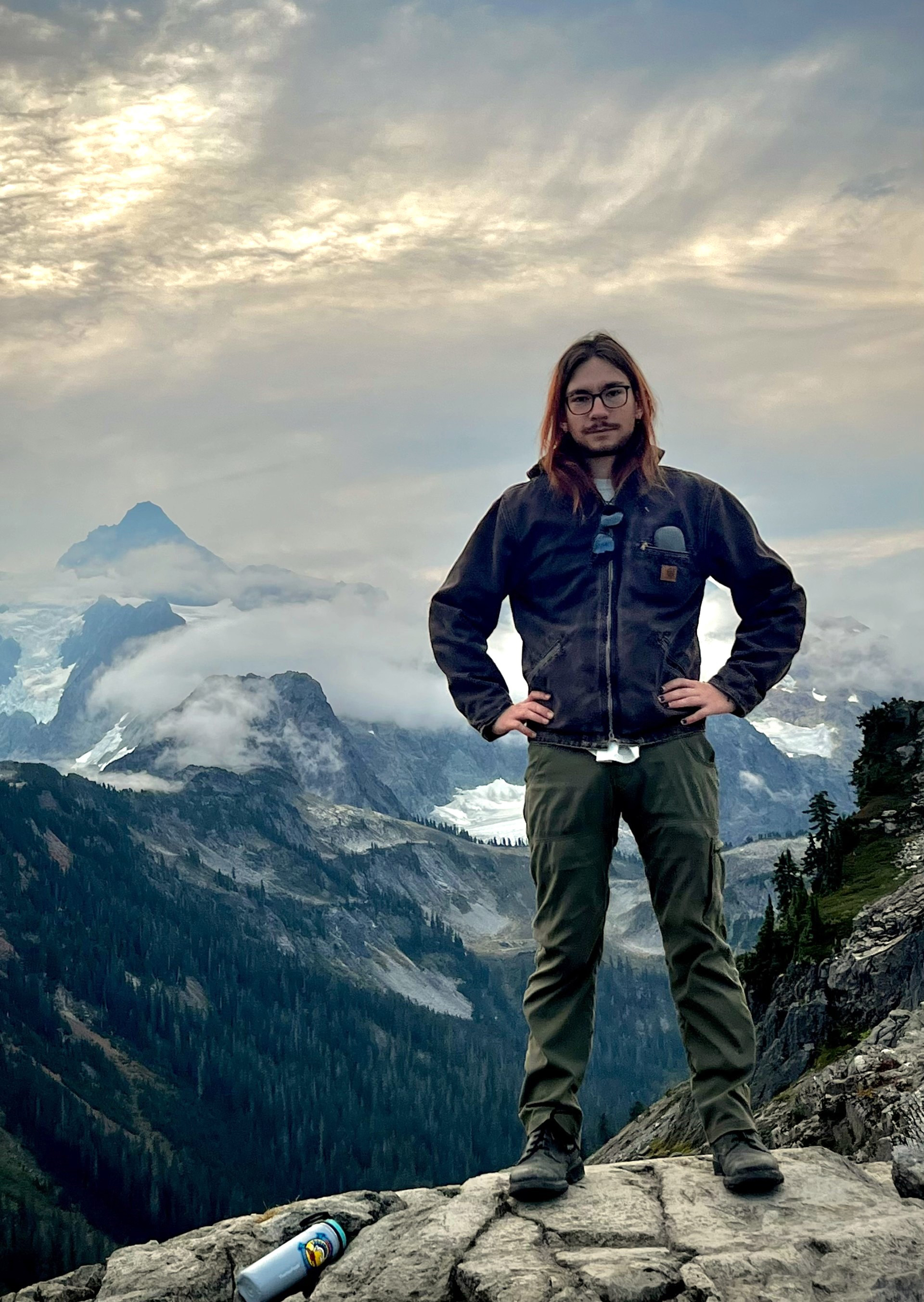 Liam KenWood stands on a rocky mountain with clouds and mountains in the background.