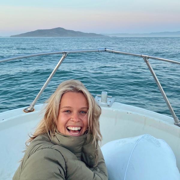 Kenna Kuhn smiles from the bow of a boat, with water and pink and blue sky behind.