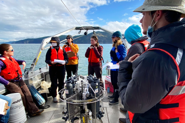 Students listen to an instructor at the stern of Western&#039;s research vessel; behind them are blue skies and one of the San Juan islands