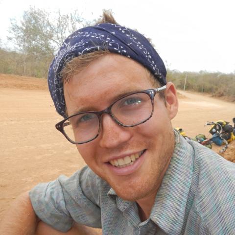 Close up portrait of Ventres-Pake smiling at the camera wearing a blue bandana as a headband and brown tortoiseshell glasses