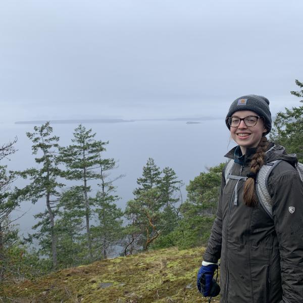 A student in a dark beanie, rain jacket, and gloves standing at the top of a mossy overlook of some thin evergreens and an overcast ocean